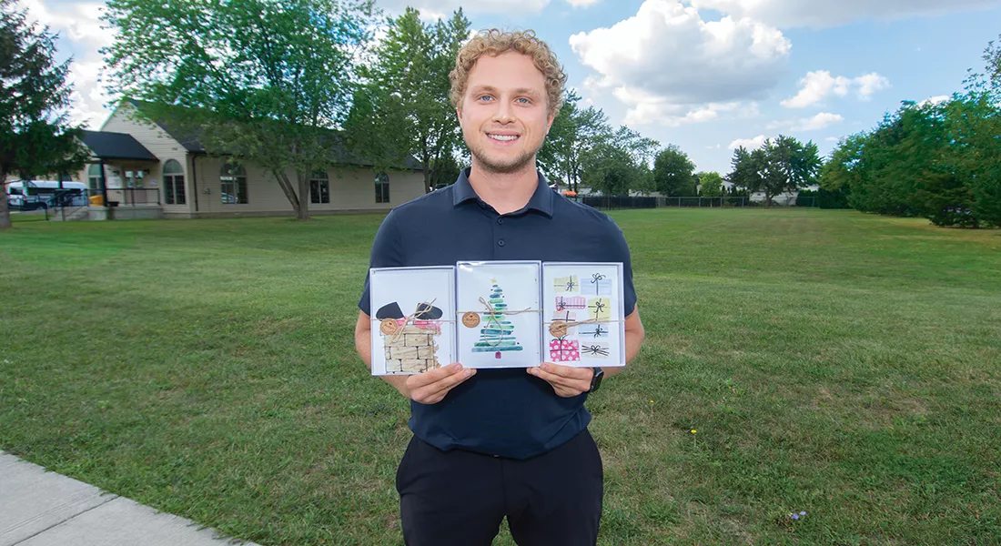 Standing on vacant land next to the Harmony in Action facilities at 3335 Woodward Blvd. in Windsor, Alexander Burrows, Harmony in Action’s Public Relations and Fundraising Coordinator, holds up this year’s selection of Christmas Cards painted by the Harmony artists. Donations from the cards and other programs will be used to build permanent residencies for individuals with disabilities, in the near future. Photo by Rod Denis - Biz X magazine